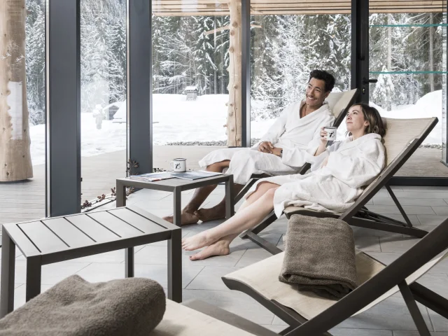 Un couple en peignoir blanc se relaxe sur des transats dans un espace bien-être de l'auberge nordique avec une vue panoramique sur une forêt enneigée.