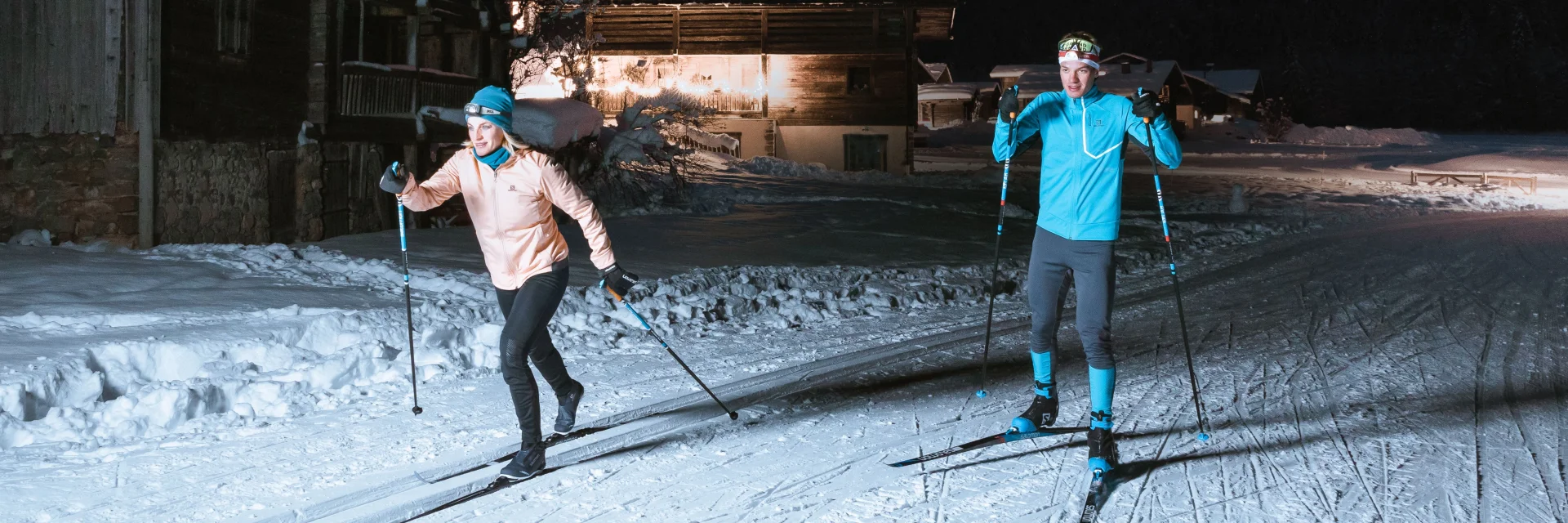 un couple de skieurs de fond sur une piste nordique du grand-bornand la nuit, ils s'éclairent à la lampe frontale et passent au travers de chalets typiques