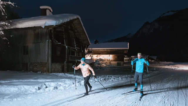 ein Paar Langläufer auf einer nordischen Loipe in grand-bornand bei Nacht, sie leuchten mit Stirnlampen und laufen durch typische Chalets