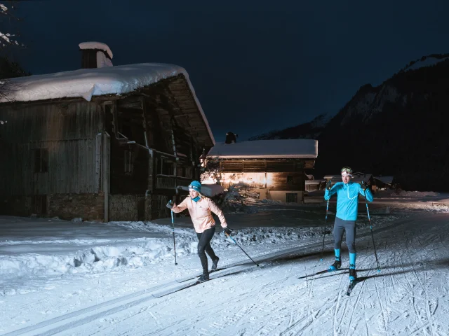 a couple of cross-country skiers on a Nordic slope in Le Grand Bornand at night, using headlamps to light their way past typical chalets