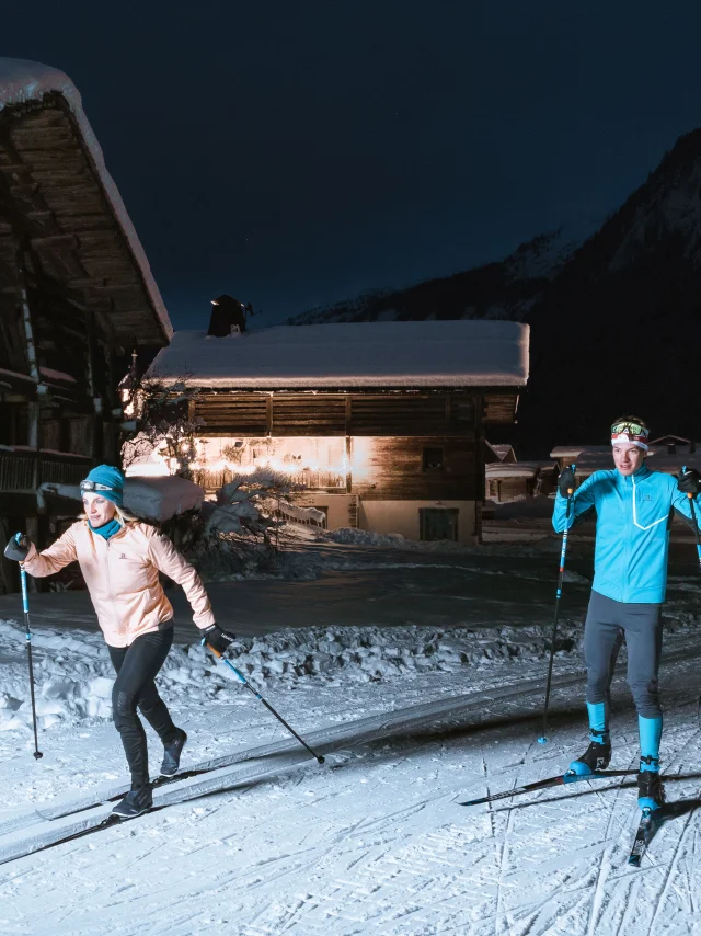 un couple de skieurs de fond sur une piste nordique du grand-bornand la nuit, ils s'éclairent à la lampe frontale et passent au travers de chalets typiques