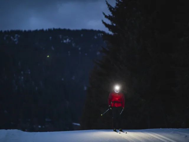 Une fondeuse de nuit sur une piste de ski nordique éclairée à la lampe frontale devant un paysage de montagne enneigée et recouverte de sapins.