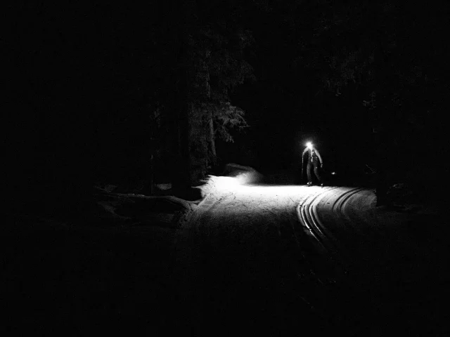 Une personne en ski de fond la nuit s'éclaire à la lampe frontale et laisse apparaitre la piste enneigée devant lui avec les traces de ski classique