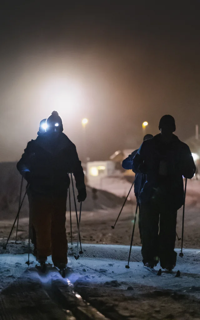 Des silhouettes de skieurs nordique en nocturne sur une piste enneigée et avec des lampes frontales