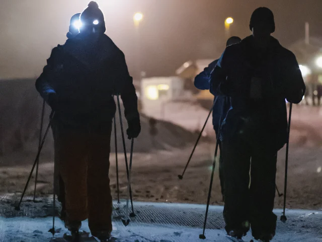 Des silhouettes de skieurs nordique en nocturne sur une piste enneigée et avec des lampes frontales