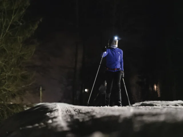 Une fondeuse équipée d'une lampe frontale fait du ski de fond classique de nuit sur une piste de ski avec des sapins l'entourant