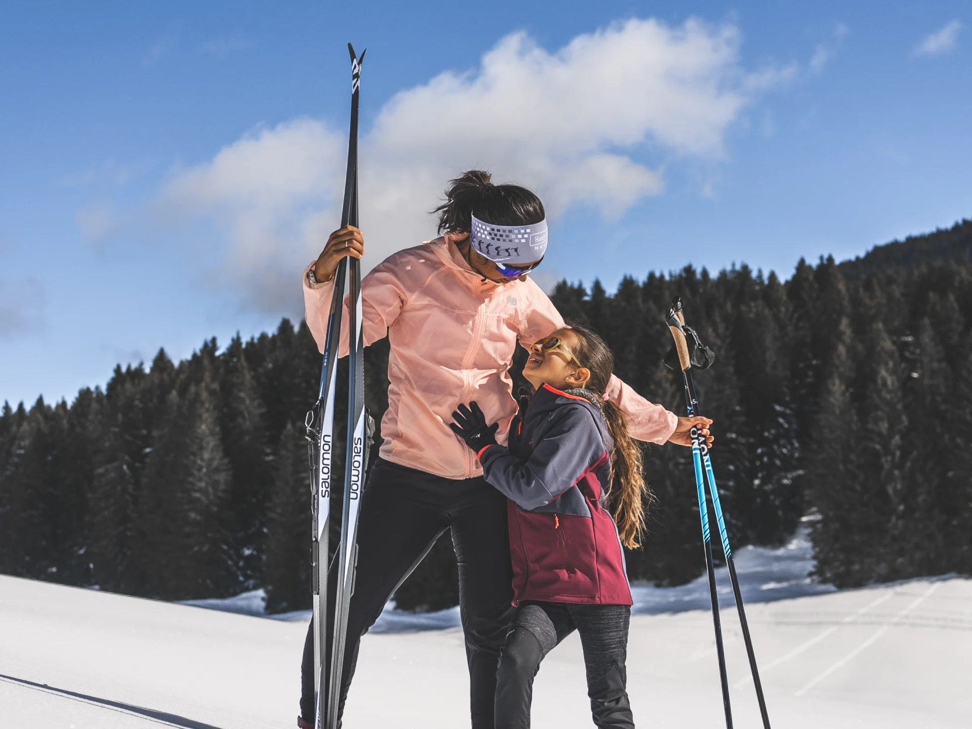Une femme et une jeune fille en tenue de ski nordique partagent un moment tendre sur une piste enneigée, sous un ciel bleu éclatant.