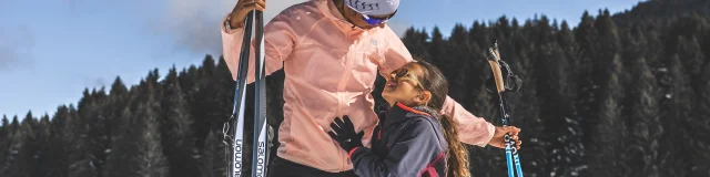 Une femme et une jeune fille en tenue de ski nordique partagent un moment tendre sur une piste enneigée, sous un ciel bleu éclatant.