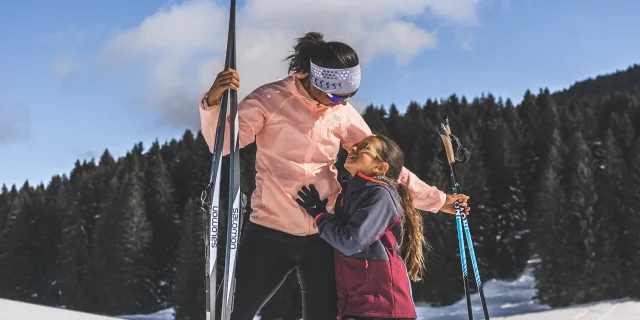 Une femme et une jeune fille en tenue de ski nordique partagent un moment tendre sur une piste enneigée, sous un ciel bleu éclatant.