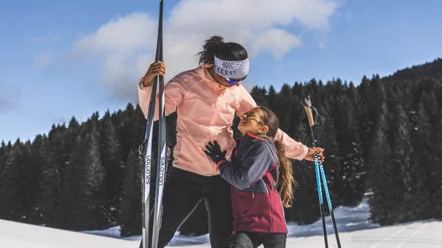 Une femme et une jeune fille en tenue de ski nordique partagent un moment tendre sur une piste enneigée, sous un ciel bleu éclatant.