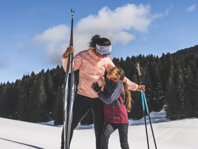 A woman and a young girl in Nordic ski gear share a tender moment on a snow-covered slope, under a brilliant blue sky.