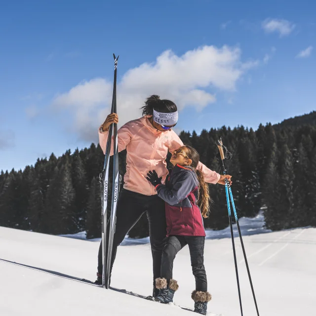 Une femme et une jeune fille en tenue de ski nordique partagent un moment tendre sur une piste enneigée, sous un ciel bleu éclatant.