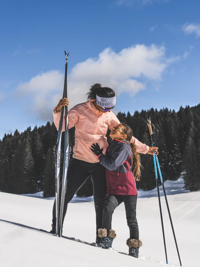 Une femme et une jeune fille en tenue de ski nordique partagent un moment tendre sur une piste enneigée, sous un ciel bleu éclatant.