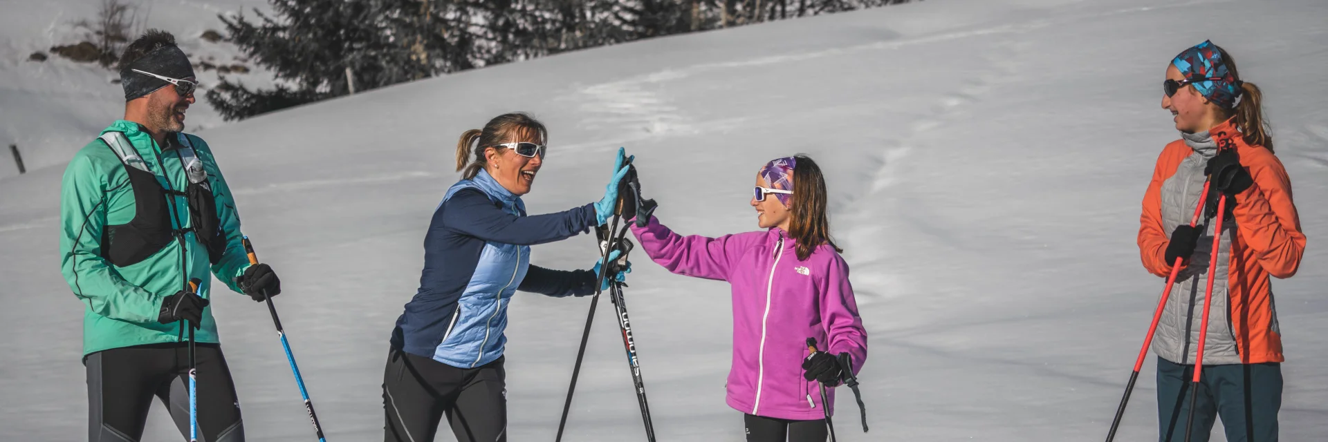 Quatre skieurs de fond en tenue colorée discutent et rient sous le soleil, tenant leurs skis et bâtons sur une piste enneigée.