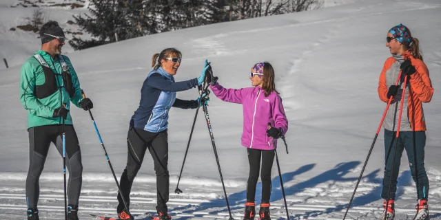 Quatre skieurs de fond en tenue colorée discutent et rient sous le soleil, tenant leurs skis et bâtons sur une piste enneigée.