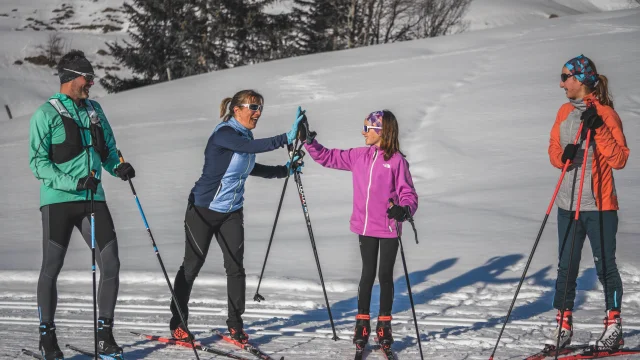Quatre skieurs de fond en tenue colorée discutent et rient sous le soleil, tenant leurs skis et bâtons sur une piste enneigée.