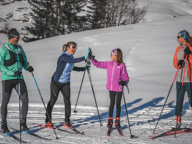 Four cross-country skiers in colourful outfits chat and laugh in the sunshine, holding their skis and poles on a snow-covered slope.
