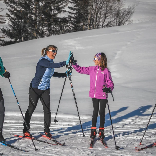 Quatre skieurs de fond en tenue colorée discutent et rient sous le soleil, tenant leurs skis et bâtons sur une piste enneigée.
