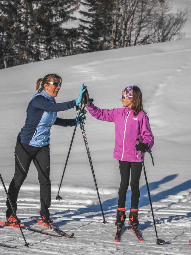 Quatre skieurs de fond en tenue colorée discutent et rient sous le soleil, tenant leurs skis et bâtons sur une piste enneigée.