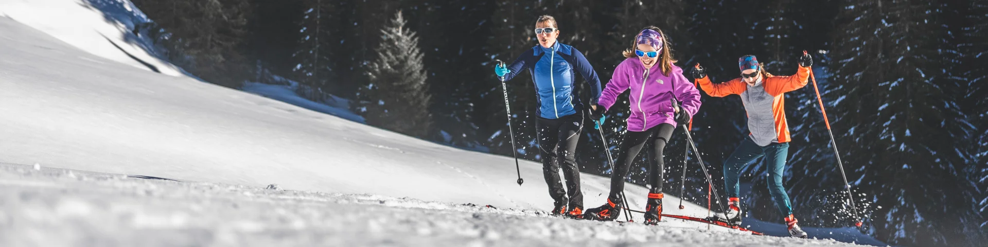 Trois skieurs en tenue colorée pratiquant le ski de fond sur un sentier enneigé, entourés de sapins et de montagnes sous un ciel bleu.
