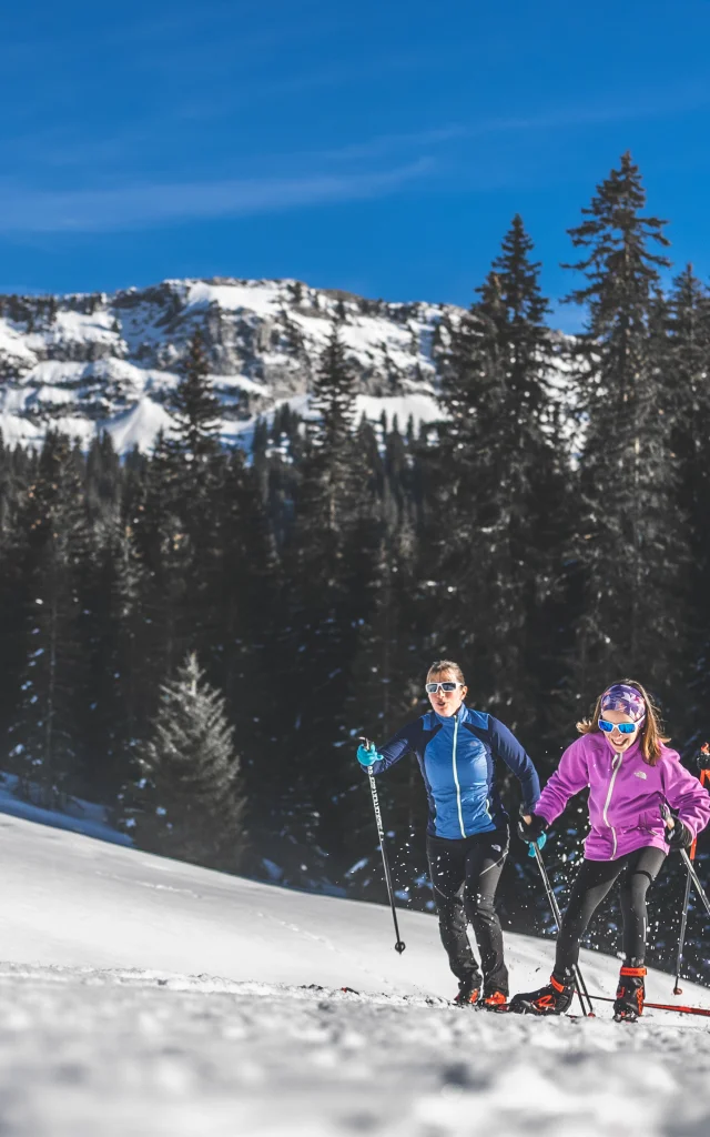 Three skiers in colourful outfits cross-country skiing on a snowy trail, surrounded by fir trees and mountains under a blue sky.