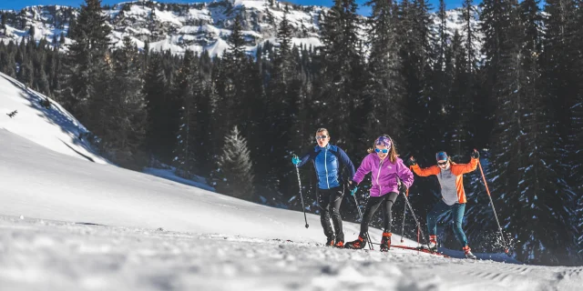 Trois skieurs en tenue colorée pratiquant le ski de fond sur un sentier enneigé, entourés de sapins et de montagnes sous un ciel bleu.