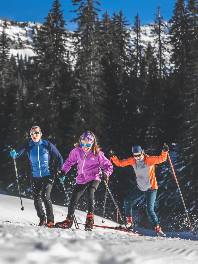Trois skieurs en tenue colorée pratiquant le ski de fond sur un sentier enneigé, entourés de sapins et de montagnes sous un ciel bleu.