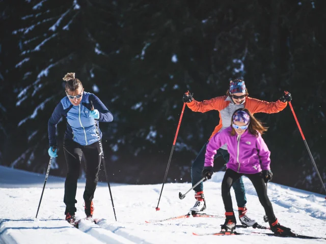 Trois skieurs en tenue colorée pratiquant le ski de fond sur un sentier enneigé, entourés de sapins et de montagnes sous un ciel bleu.