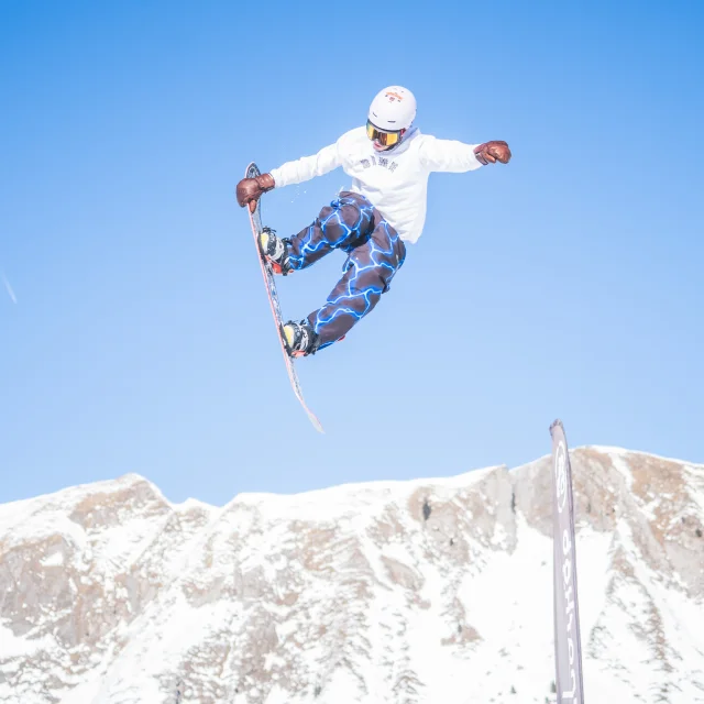 Un snowboardeur est dans les airs et attrape sa planche avec sa main droite, en arrière plan se trouve l'arrête de la pointe d'almet