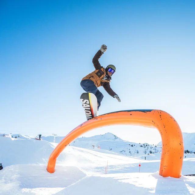 Un snowboardeur glisse sur un rainbow rail sous un ciel dégagé avec en arrière plan les crêtes du maroly et le mont lachat