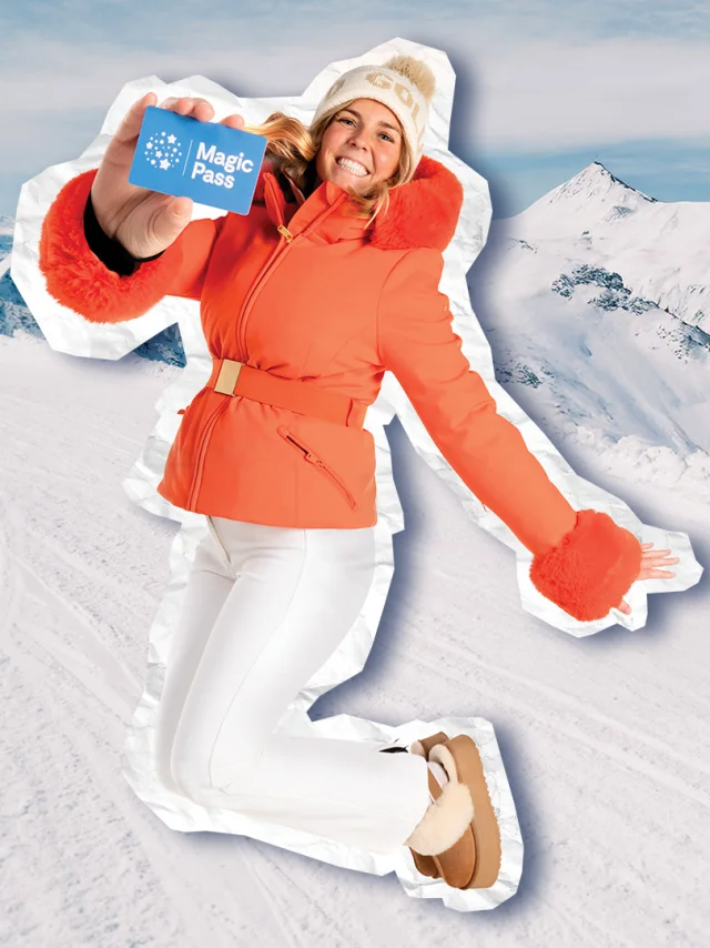 Une femme souriante, vêtue d’une veste orange et d’un pantalon blanc, saute de joie en tenant un Magic Pass, avec un panorama de montagnes enneigées en arrière-plan.