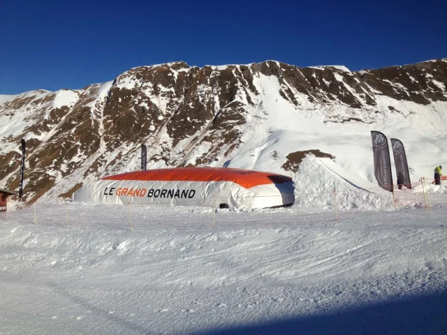 Un grand coussin gonflable blanc et orange avec l'inscription Le Grand Bornand, installé sur une piste enneigée au pied de montagnes rocheuses partiellement recouvertes de neige.