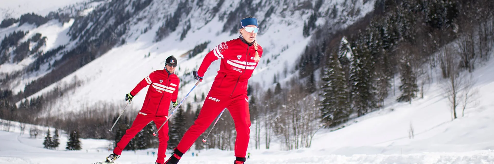 Deux moniteurs nordiques de l'école de ski Français se suivent en skating sur une piste enneigée entourée d'un paysage montagnard enneigé et rempli de sapins