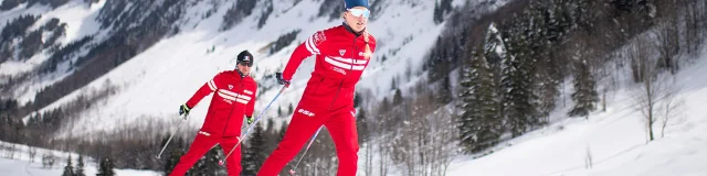 Deux moniteurs nordiques de l'école de ski Français se suivent en skating sur une piste enneigée entourée d'un paysage montagnard enneigé et rempli de sapins
