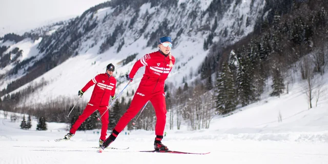 Deux moniteurs nordiques de l'école de ski Français se suivent en skating sur une piste enneigée entourée d'un paysage montagnard enneigé et rempli de sapins