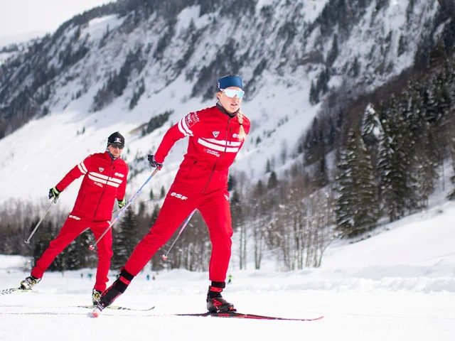 Zwei Nordic-Lehrer der französischen Skischule folgen einander im Skating auf einer verschneiten Piste, die von einer verschneiten Berglandschaft voller Tannen umgeben ist.