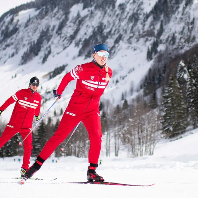 Deux moniteurs nordiques de l'école de ski Français se suivent en skating sur une piste enneigée entourée d'un paysage montagnard enneigé et rempli de sapins