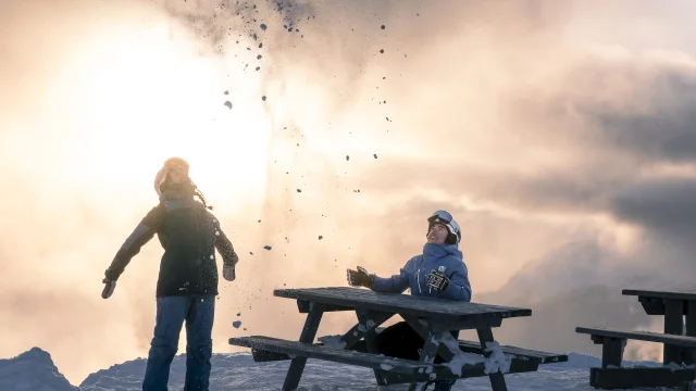 Deux skieurs en tenue d’hiver s’amusent dans la neige au sommet d’une montagne, l’un lançant de la neige en l’air, l’autre assis à une table et souriant, avec un coucher de soleil en toile de fond.