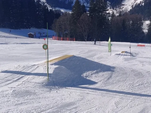 Un snowpark en montagne avec des modules pour débutants sur une piste de ski, entouré d'arbres et de montagnes enneigées sous un ciel ensoleillé.