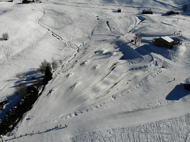 Photo prise par drone montrant un snowpark sur une pente enneigée avec plusieurs modules de saut, des bâtiments de montagne et des skieurs en file indienne en contrebas.