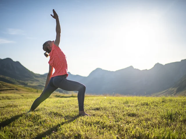 Une femme pratiquant le yoga dans un champ verdoyant de montagne, en pleine posture du guerrier, sous un soleil éclatant.