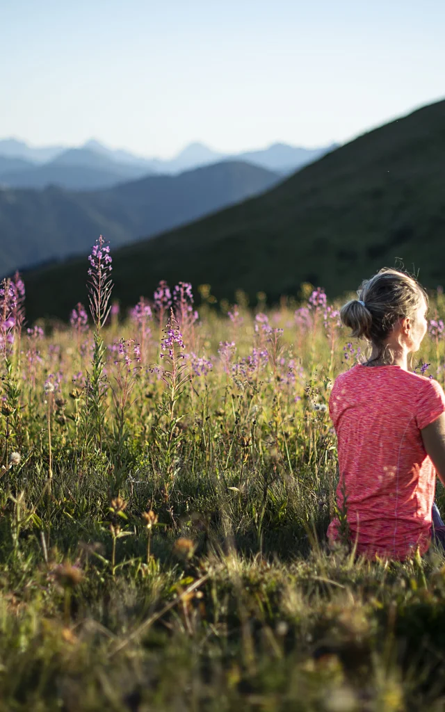 A woman practising yoga in a green mountain field, in the lotus posture, surrounded by violet flowers.