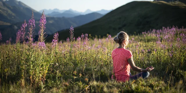Une femme pratiquant le yoga dans un champ verdoyant de montagne, en pleine posture du lotus, au milieu de fleurs de couleurs violette