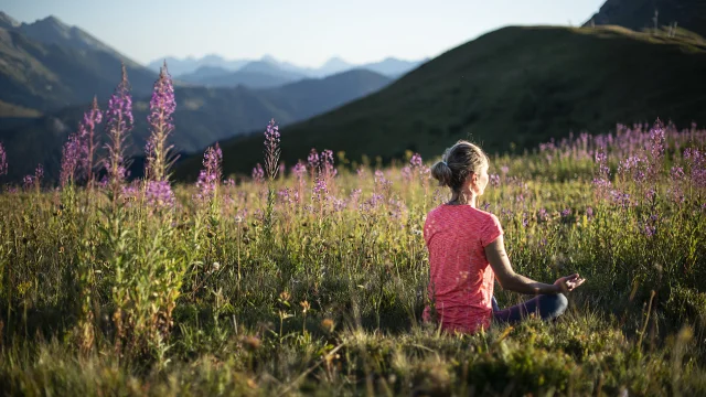 Une femme pratiquant le yoga dans un champ verdoyant de montagne, en pleine posture du lotus, au milieu de fleurs de couleurs violette