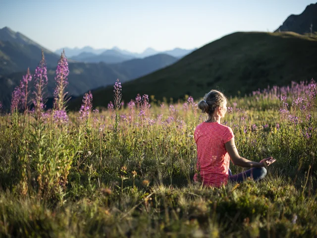 Une femme pratiquant le yoga dans un champ verdoyant de montagne, en pleine posture du lotus, au milieu de fleurs de couleurs violette