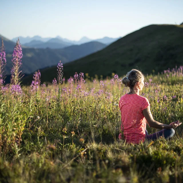 Une femme pratiquant le yoga dans un champ verdoyant de montagne, en pleine posture du lotus, au milieu de fleurs de couleurs violette