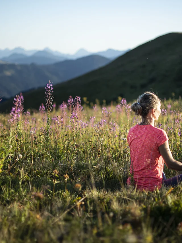 Une femme pratiquant le yoga dans un champ verdoyant de montagne, en pleine posture du lotus, au milieu de fleurs de couleurs violette