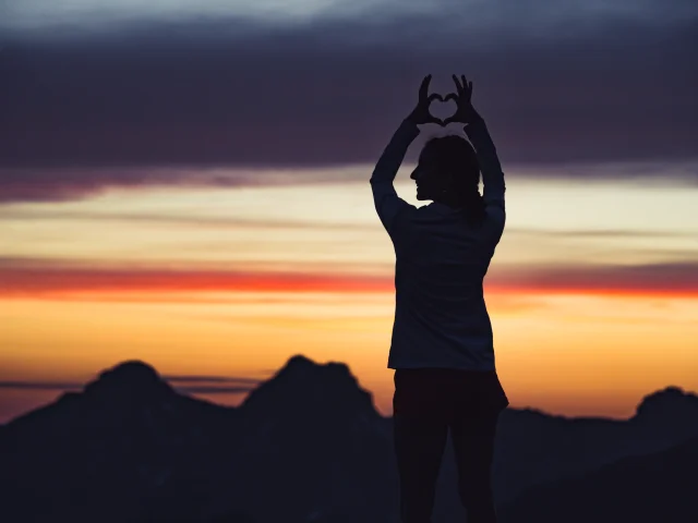 Une silhouette effectuant un coeur avec ses mains sur un sommet montagneux, avec un coucher de soleil éclatant en arrière-plan.