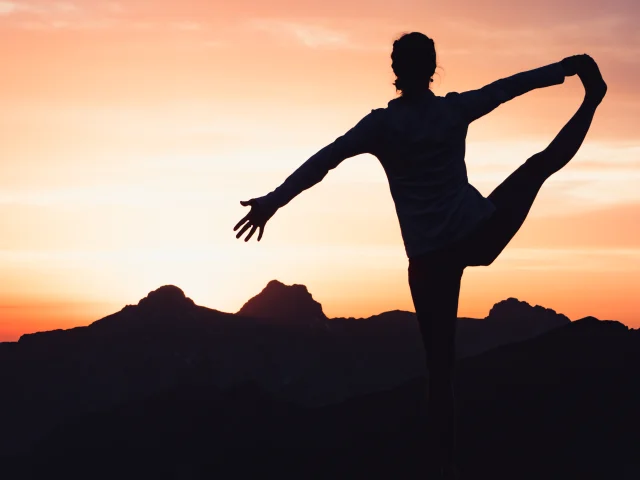 Une silhouette effectuant une posture de yoga sur un sommet montagneux, avec un coucher de soleil éclatant en arrière-plan.