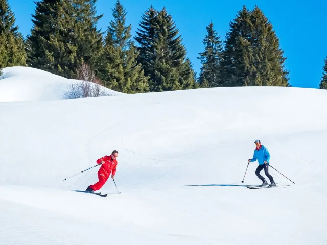 Deux skieurs de fond glissent sur une vaste étendue de neige fraîche, entourés de sapins et sous un ciel bleu éclatant.